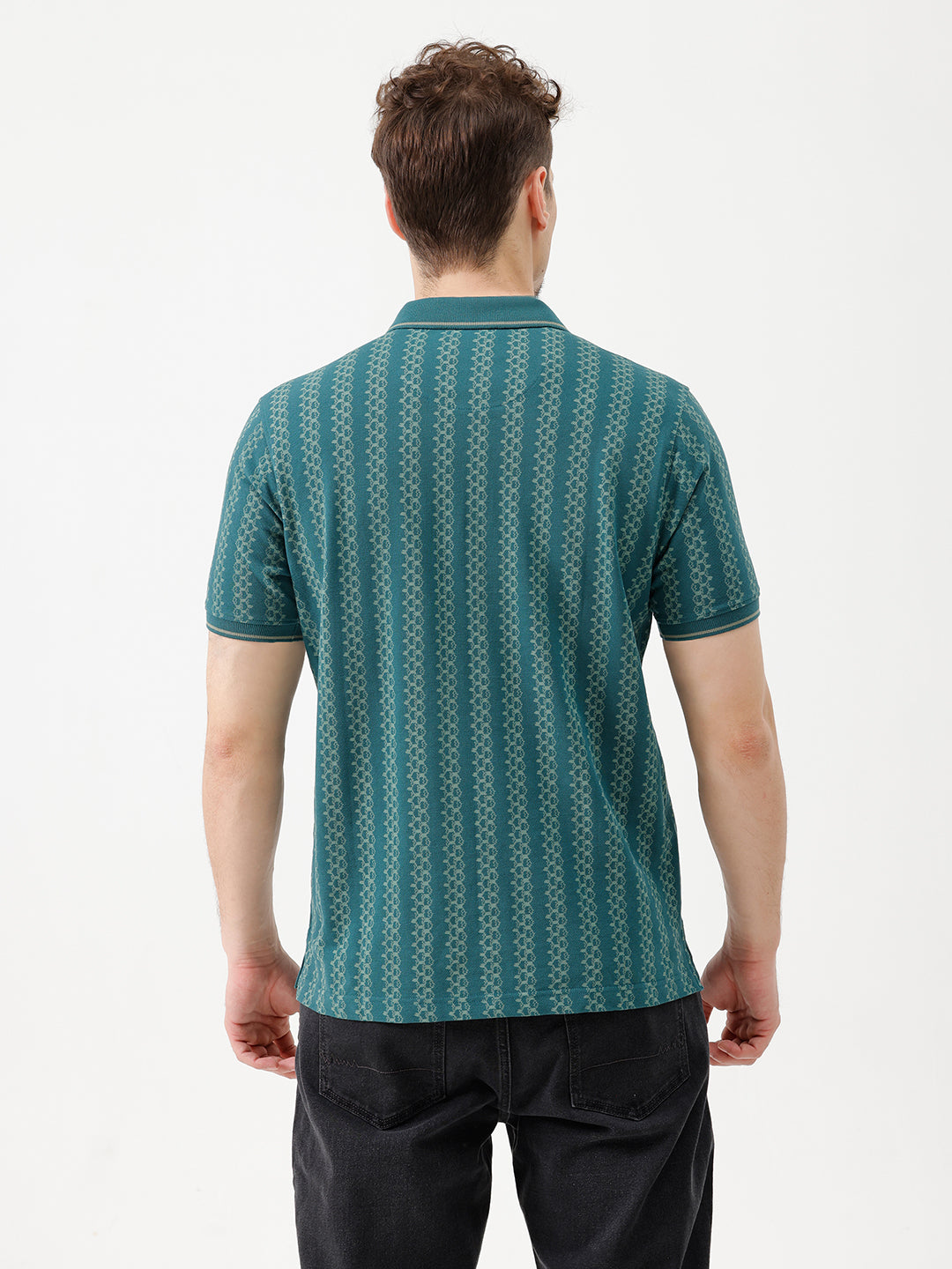 A man wearing a teal green short-sleeve polo shirt with a white geometric pattern, paired with black jeans, standing against a plain white studio background.