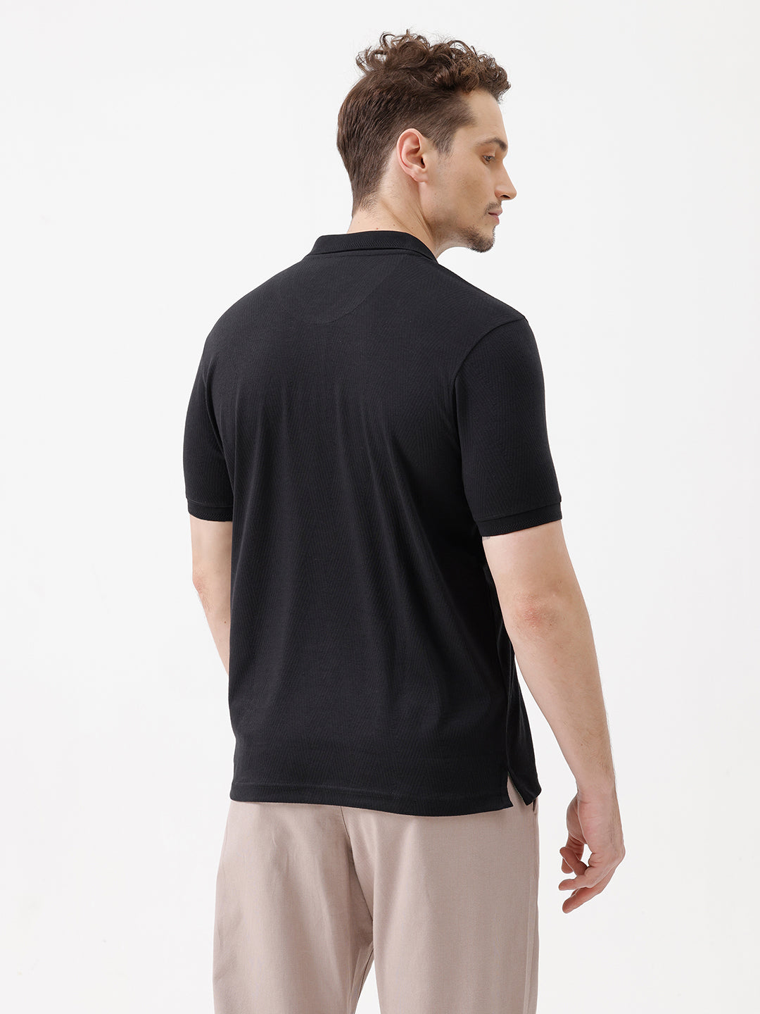 A man wearing a black short-sleeve polo shirt with a single chest pocket, paired with beige trousers, standing against a plain white studio background.