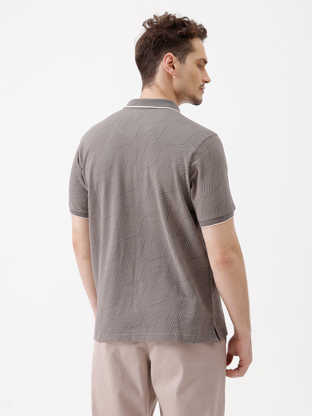 A man wearing a short-sleeve polo shirt with a gray and white geometric pattern, paired with beige trousers, standing against a plain white studio background.