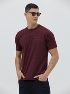 A man wearing a short-sleeve crewneck t-shirt in maroon with a subtle tonal graphic pattern, paired with dark blue jeans, against a light gray studio background.