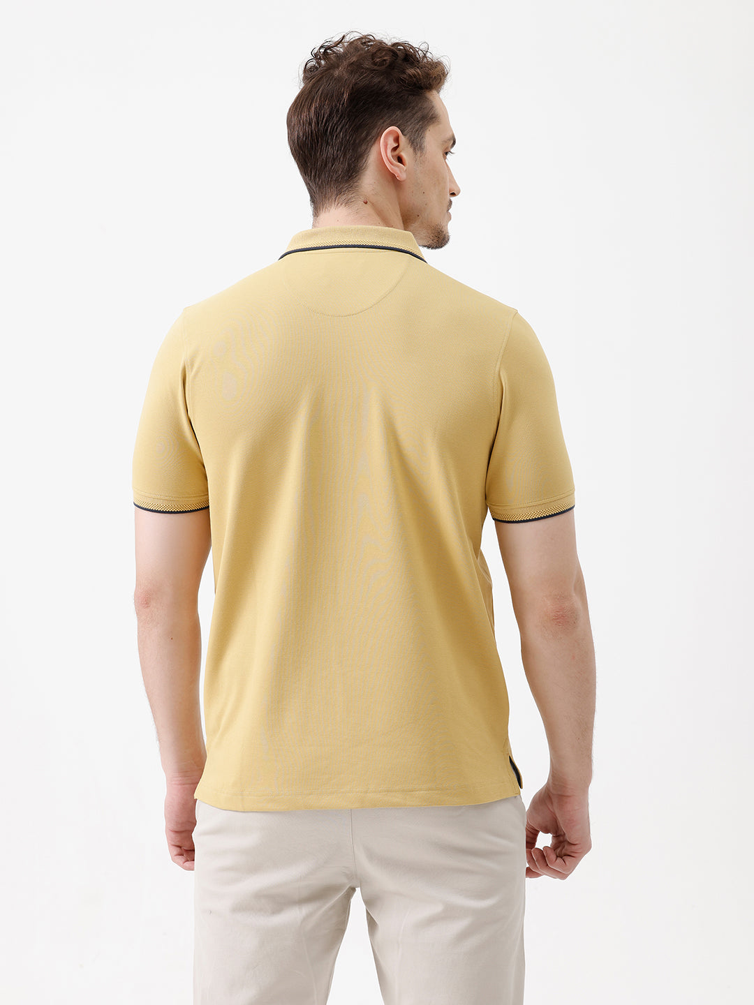 A man wearing a light yellow Lacoste polo shirt with dark piping on the collar and sleeve cuffs, paired with beige trousers, against a plain white studio background.