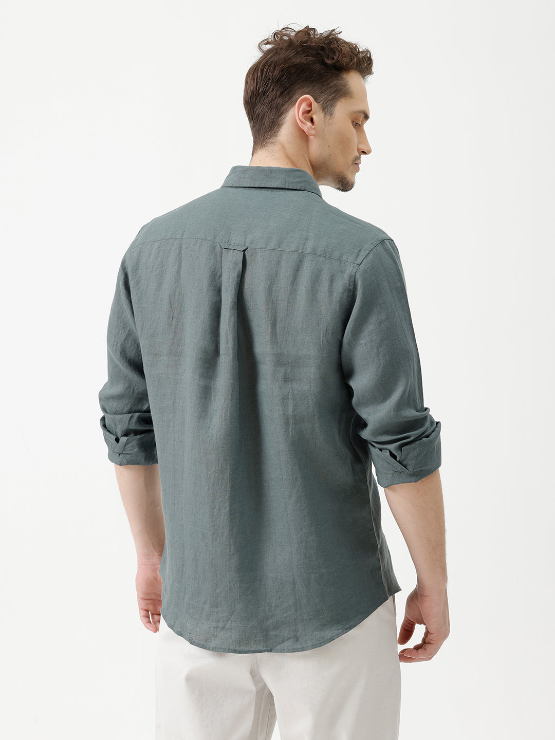 A man wearing a slate blue-green long-sleeve linen button-up shirt with white trousers, standing against a plain white studio background.