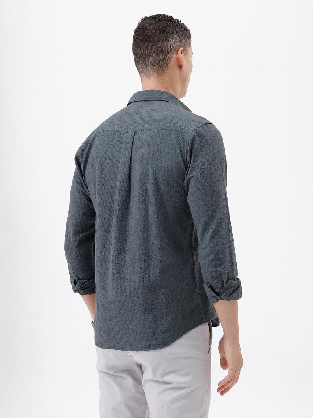 A man wearing a dark charcoal gray long-sleeve button-down shirt with a chest pocket, paired with light gray trousers, standing against a plain white studio background.