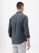 A man wearing a dark charcoal gray long-sleeve button-down shirt with a chest pocket, paired with light gray trousers, standing against a plain white studio background.