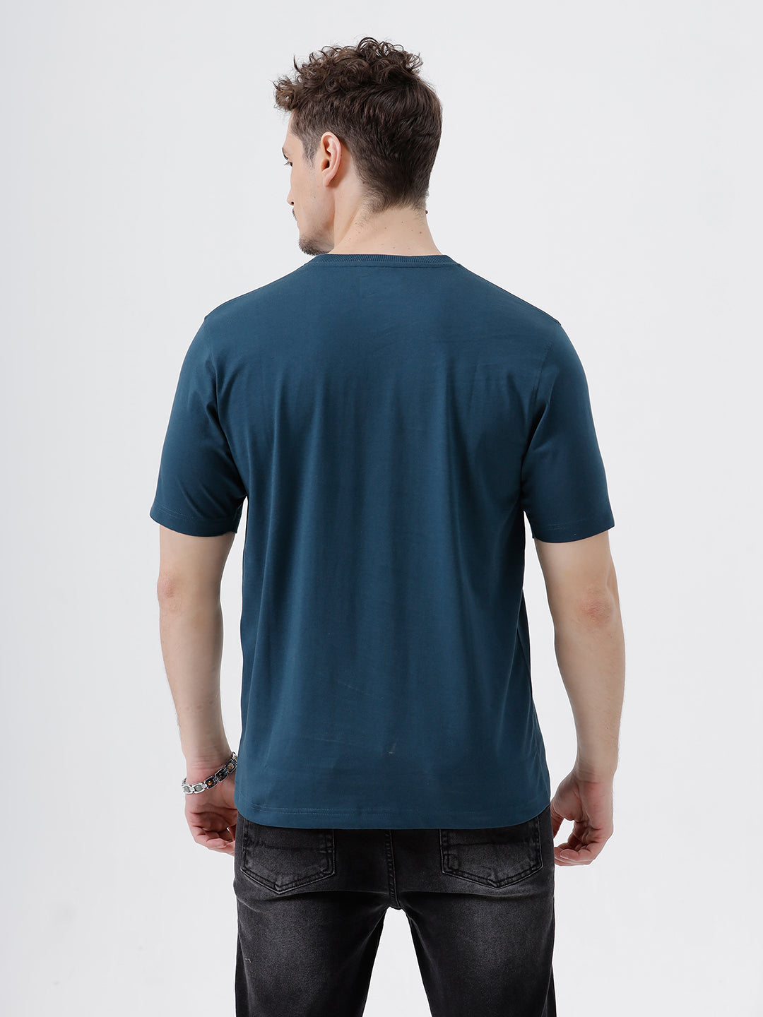 A man wearing a dark navy blue crew-neck t-shirt and black jeans, standing against a light gray studio background.