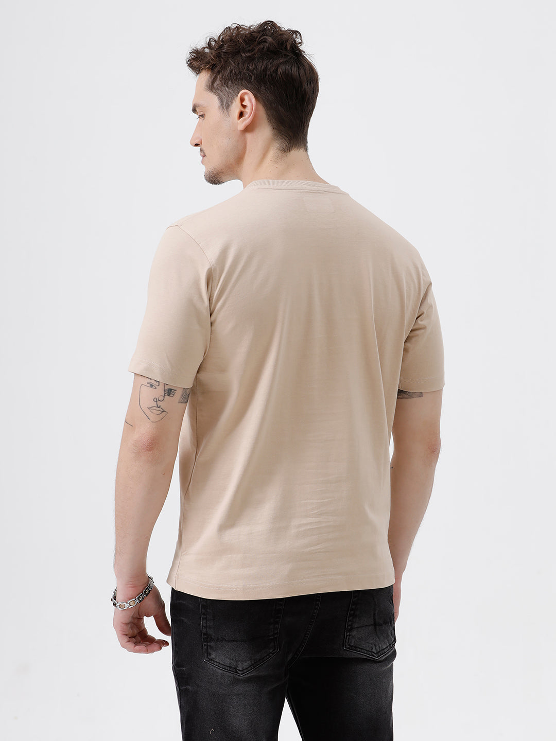 A man wearing a beige short-sleeve crewneck t-shirt with a subtle tonal logo on the left chest, paired with black jeans, standing against a light gray studio background.