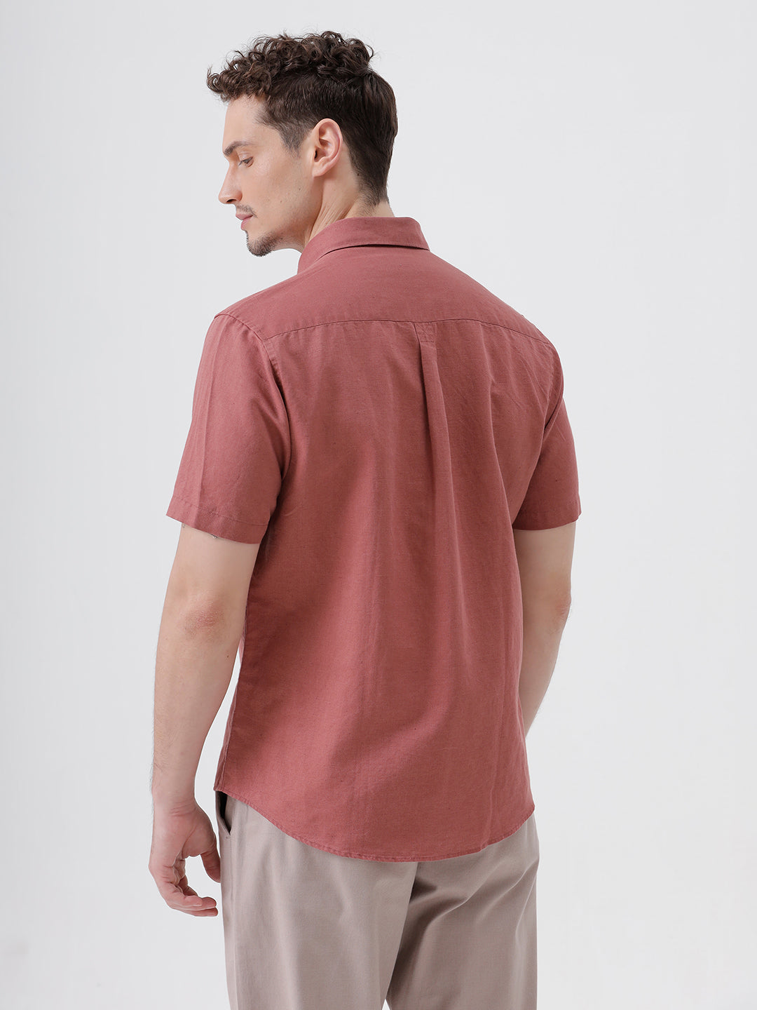 A man wearing a short-sleeve button-up shirt in a dusty rose color, paired with beige trousers, standing against a light gray studio background.