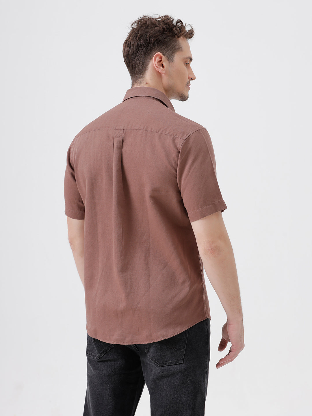 A man wearing a short-sleeve button-up shirt in a dusty rose-brown color, paired with black jeans, standing against a plain light gray background.