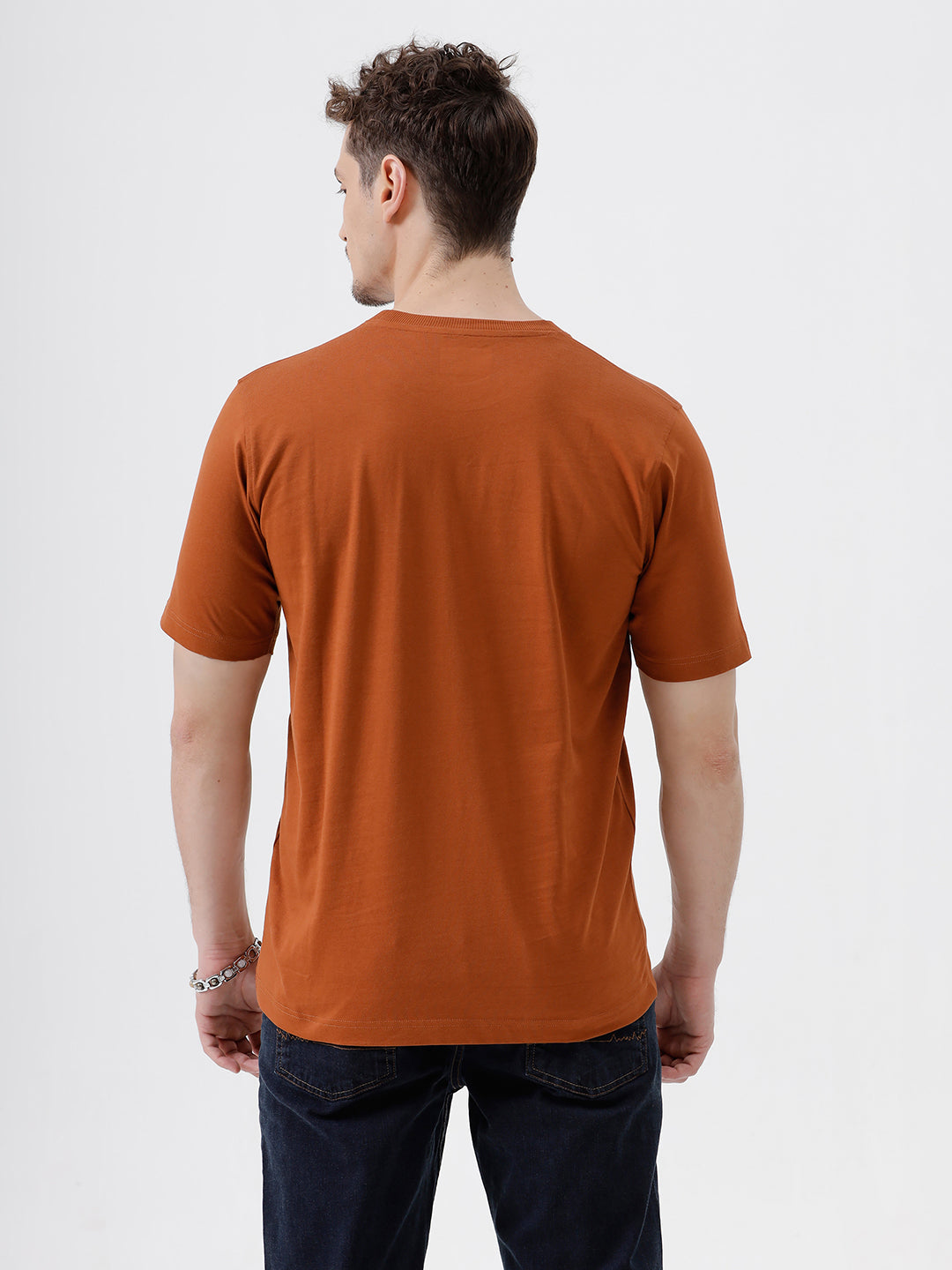 A man wearing a rust-orange crew-neck t-shirt and dark blue jeans, standing against a light gray studio background.