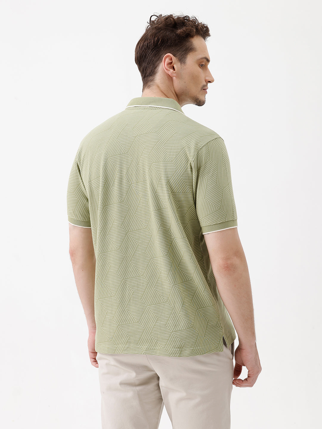 A man wearing a light sage green short-sleeve polo shirt with a subtle geometric pattern, paired with beige trousers, standing against a plain white studio background.