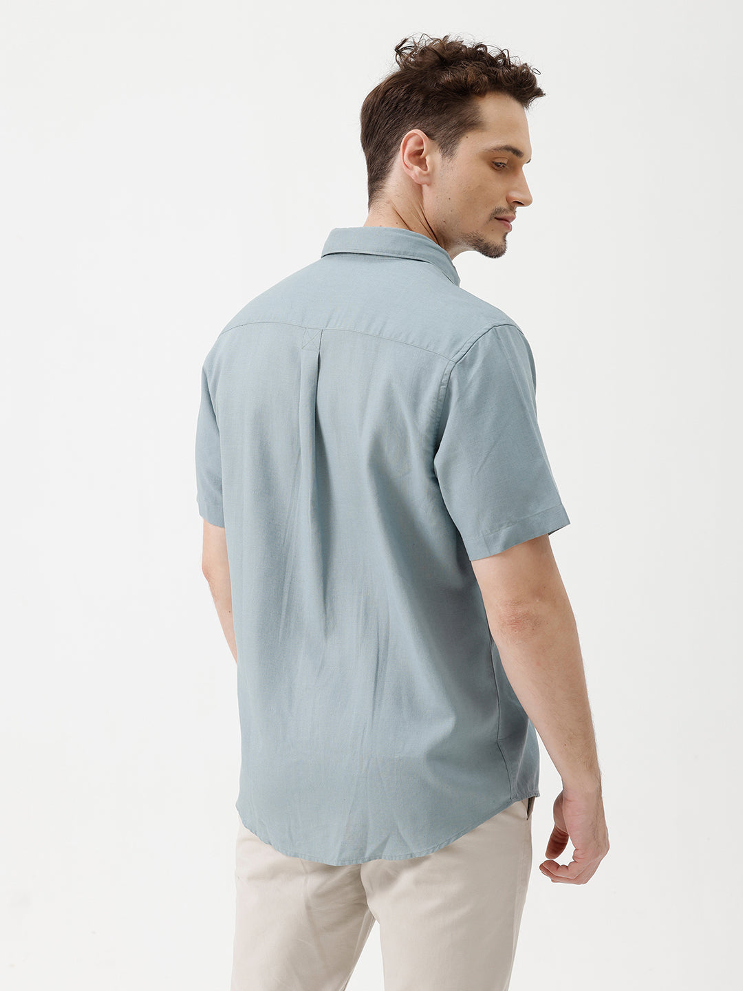 A man wearing a light blue short-sleeve button-up shirt and beige trousers, standing against a plain white studio background.