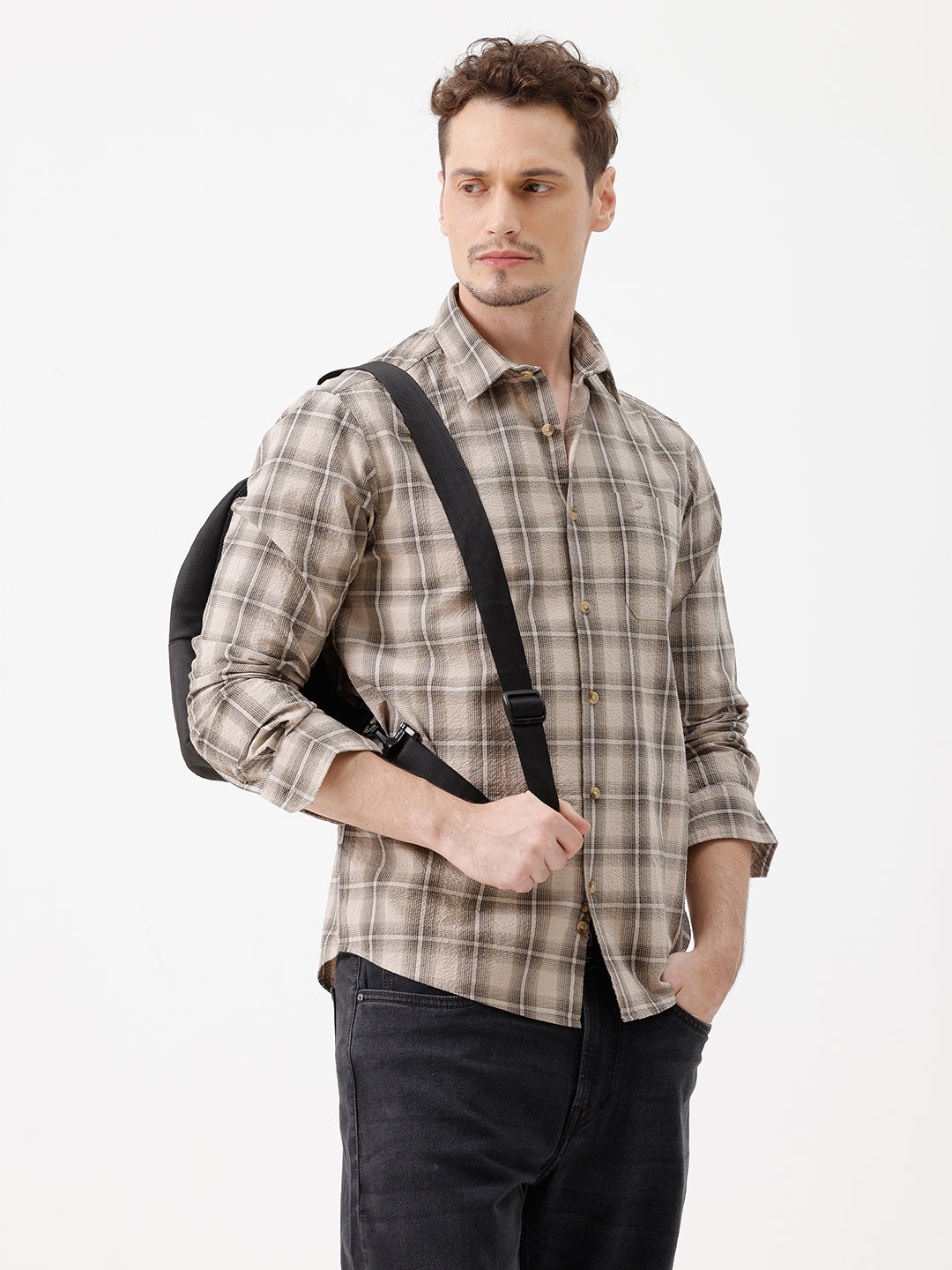 A man wearing a long-sleeve button-up plaid shirt in beige and brown tones, paired with dark jeans, standing against a plain white studio background.