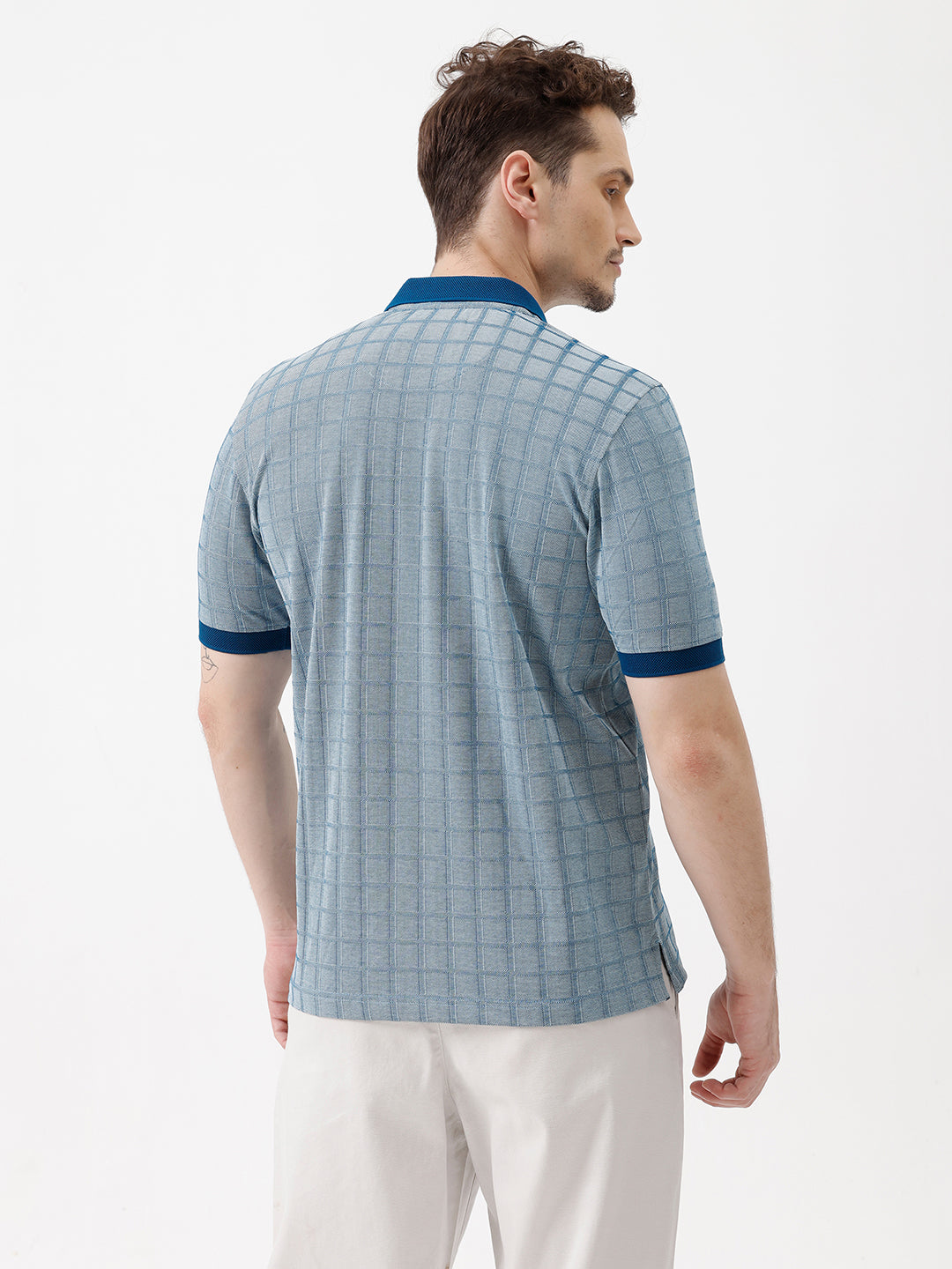 A man wearing a light blue and white checkered polo shirt with a dark blue collar and sleeve cuffs, paired with white trousers, standing against a plain white background.