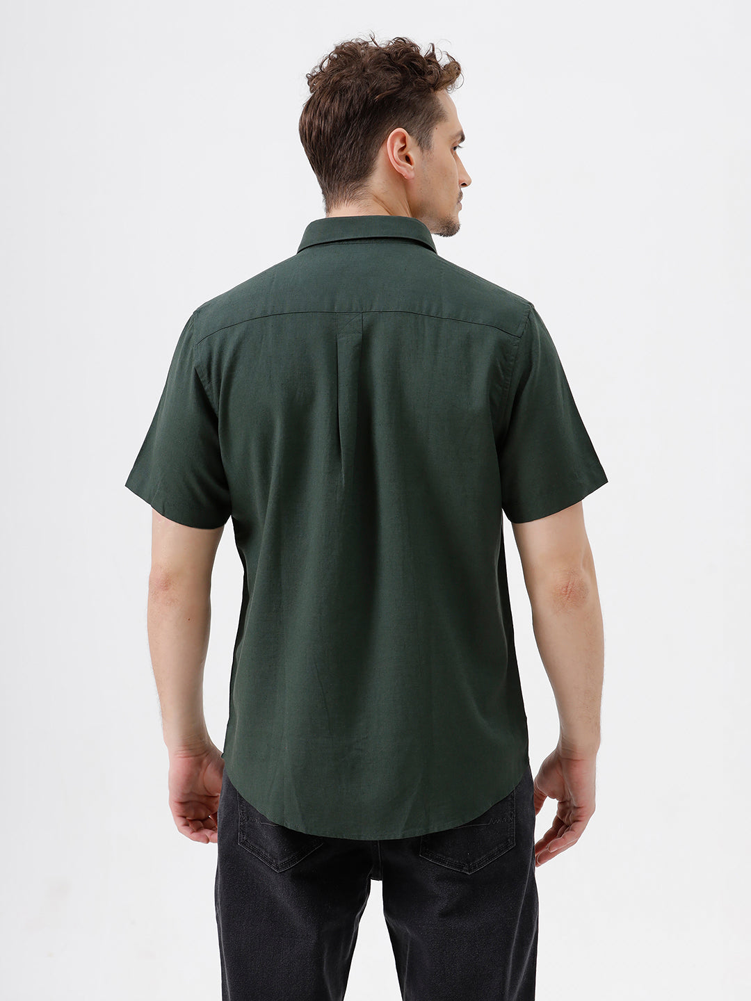 A man wearing a dark forest green short-sleeve button-up shirt with a chest pocket, paired with black jeans, standing against a plain white studio background.