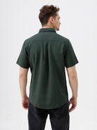 A man wearing a dark forest green short-sleeve button-up shirt with a chest pocket, paired with black jeans, standing against a plain white studio background.