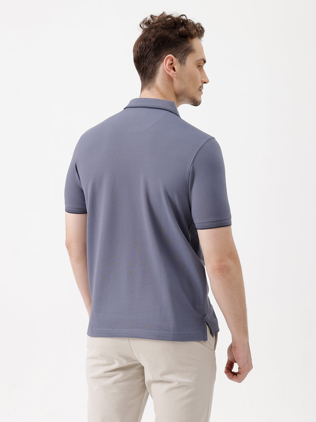 A man wearing a slate blue polo shirt and beige trousers, standing against a plain white background.