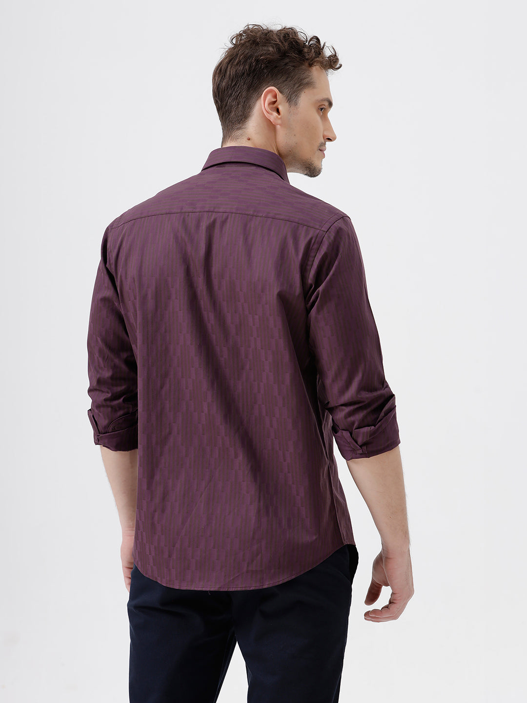 A man wearing a dark purple long-sleeve button-down shirt with subtle vertical stripes and dark navy trousers, standing against a light gray background.