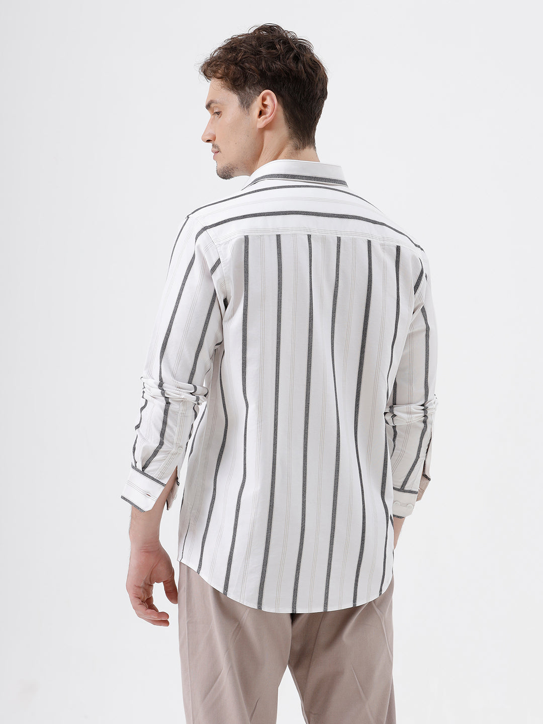 A man wearing a white long-sleeve button-up shirt with vertical gray stripes and beige trousers, standing against a light gray studio background.