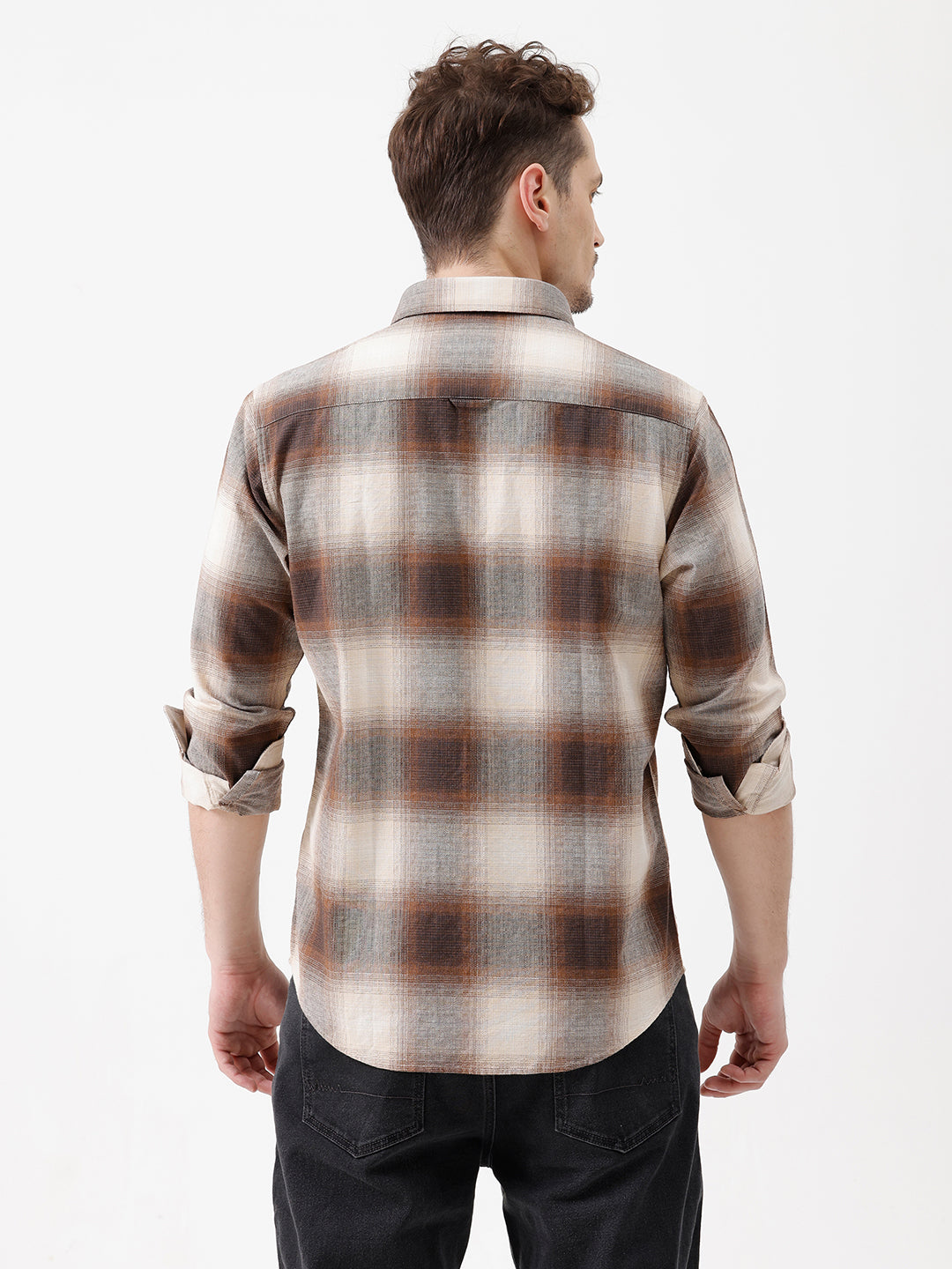 A man wearing a long-sleeve button-up plaid shirt in brown, beige, and gray tones, paired with dark jeans, standing against a plain white studio background.