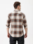 A man wearing a long-sleeve button-up plaid shirt in brown, beige, and gray tones, paired with dark jeans, standing against a plain white studio background.