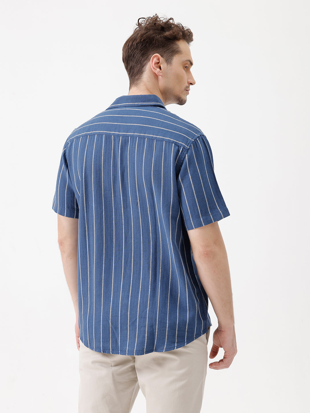 A man wearing a blue short-sleeve button-up shirt with thin white vertical stripes and beige trousers, standing against a plain white studio background.