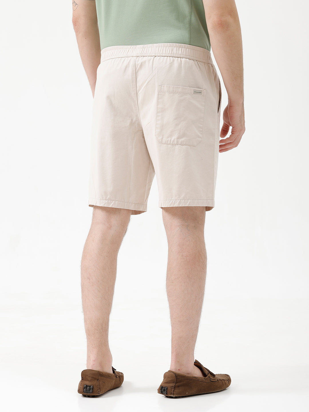 A man wearing light beige chino shorts, a sage green t-shirt, and brown suede loafers, shown from the waist down against a white studio background.