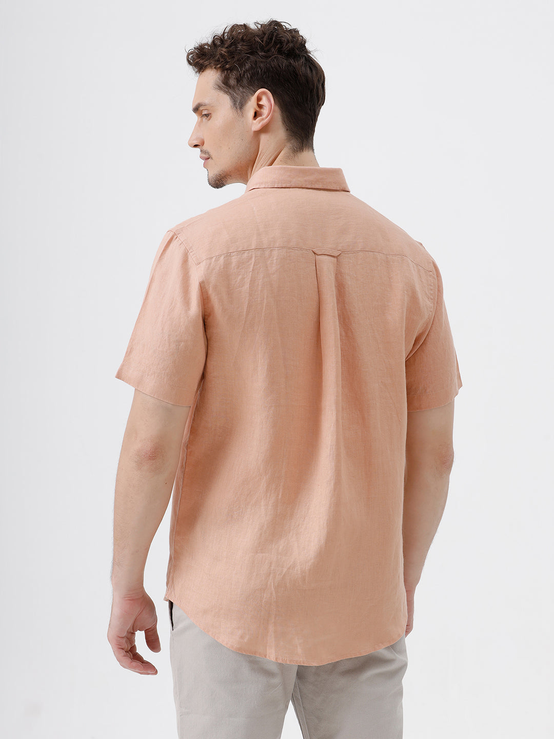 A man wearing a short-sleeve linen button-up shirt in a light peach-pink color, paired with light gray trousers, standing against a plain light gray background.