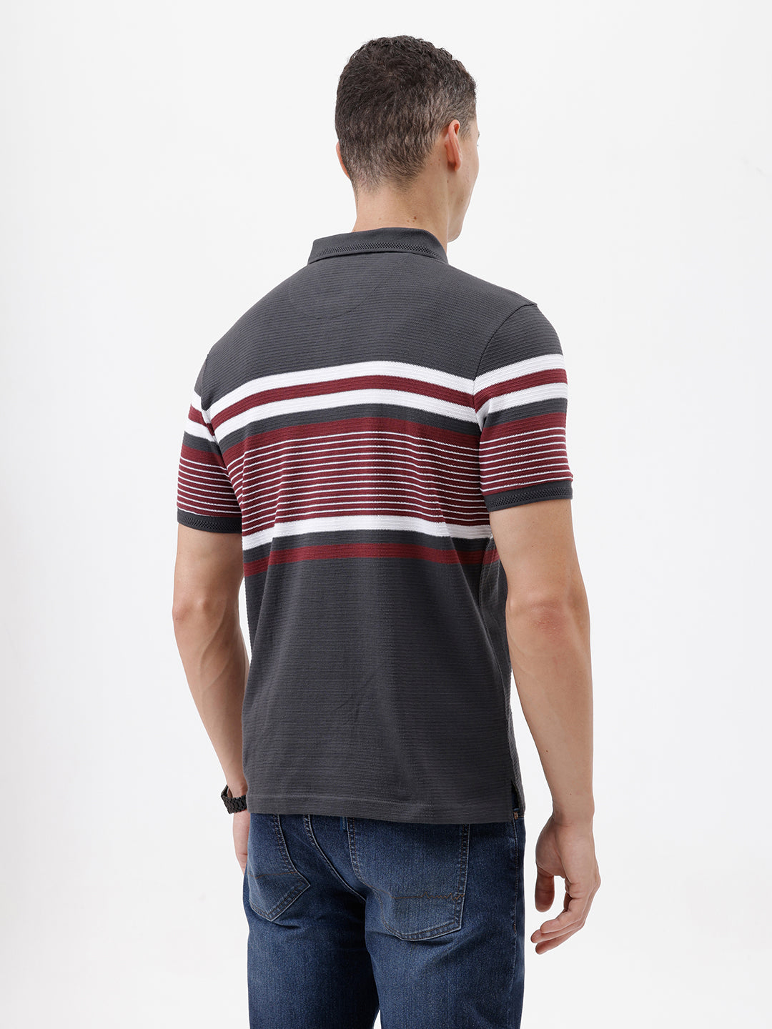 A man wearing a dark gray Lacoste polo shirt with horizontal red and white stripes across the chest and sleeves, paired with blue jeans, against a plain light gray background.