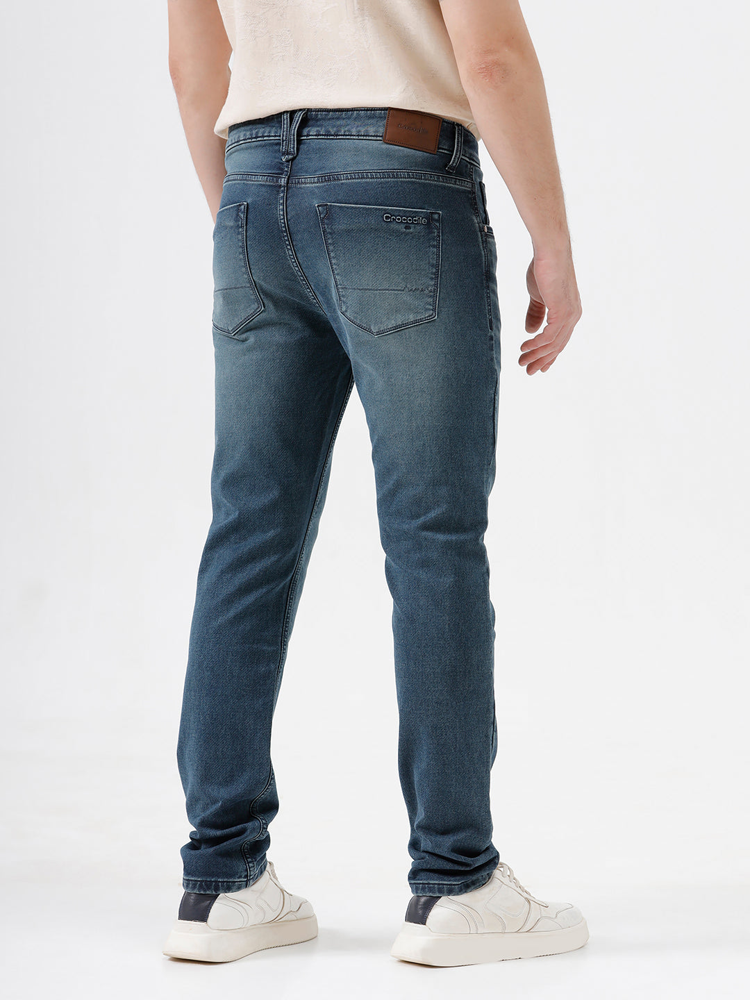 A person wearing slim-fit medium wash blue jeans, a cream-colored short-sleeve button-up shirt, and white sneakers, standing against a plain white studio background.
