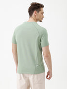 A man wearing a light sage green athletic t-shirt with a small tonal logo on the chest, paired with beige pants, standing against a plain white studio background.