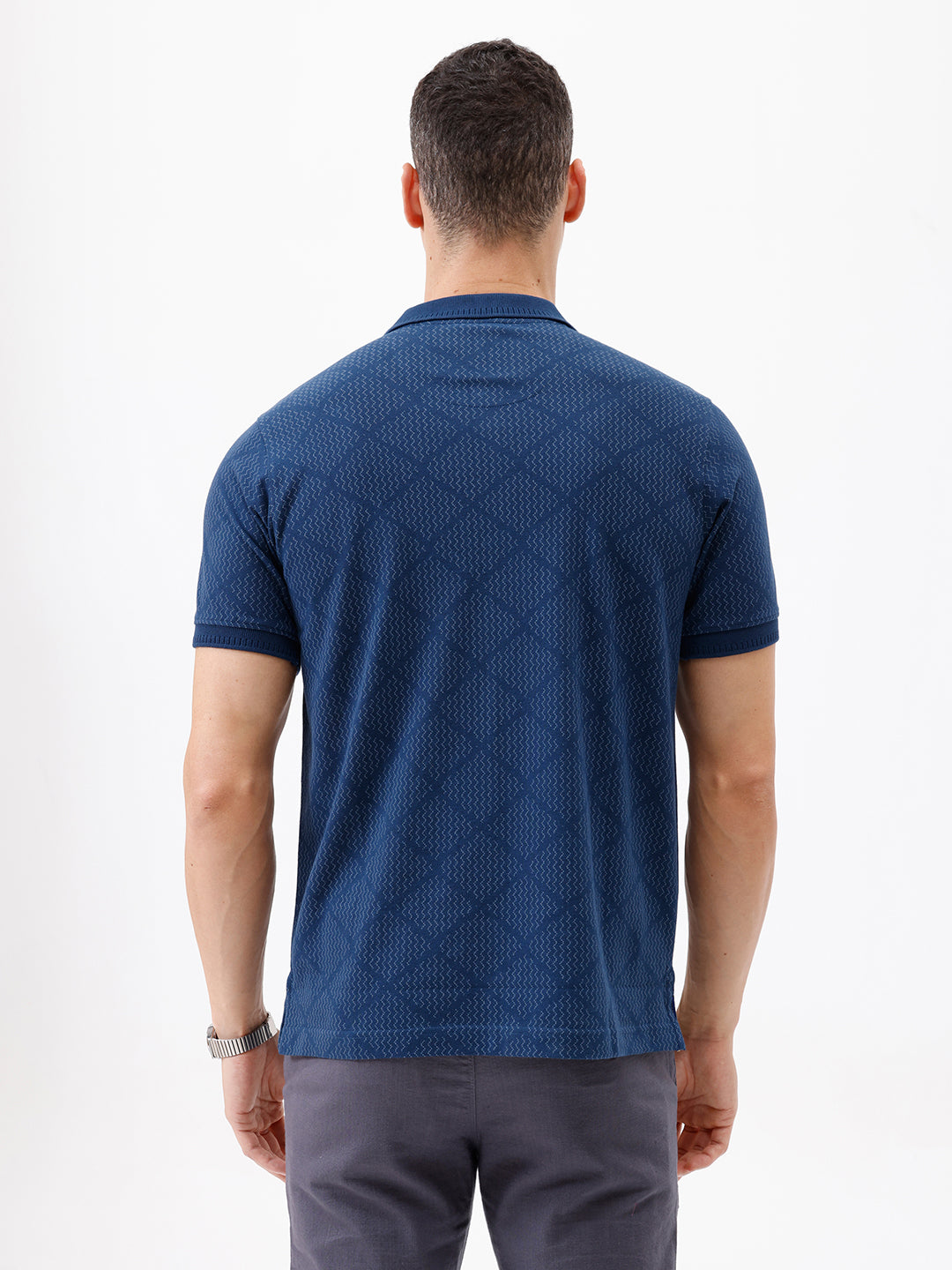 A man wearing a navy blue short-sleeve polo shirt with a subtle geometric pattern and a half-zip collar, paired with gray trousers, against a plain white background.