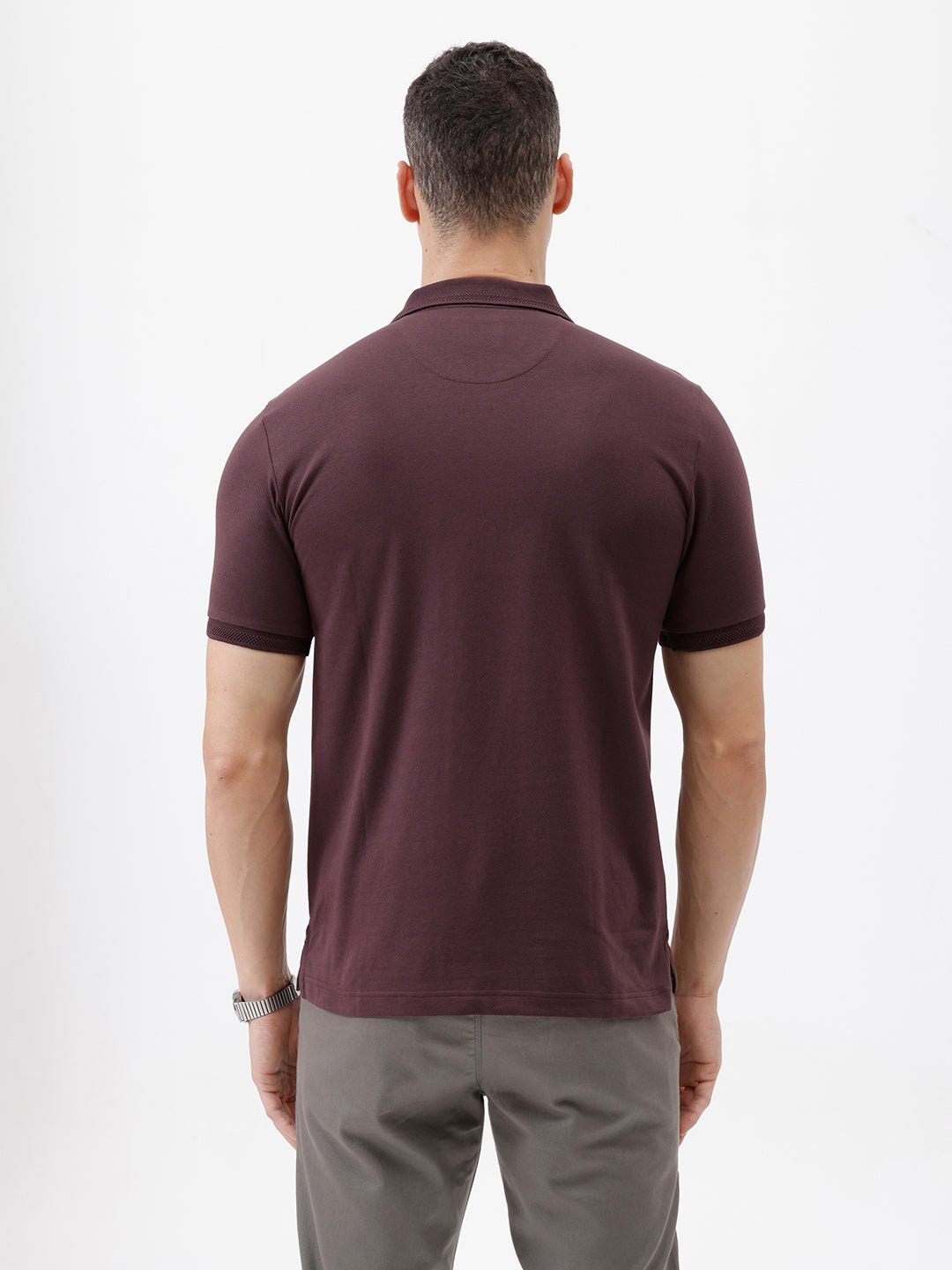 A man wearing a short-sleeve polo shirt in dark burgundy with a chest pocket, paired with gray trousers, standing against a plain light gray background.