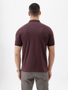 A man wearing a short-sleeve polo shirt in dark burgundy with a chest pocket, paired with gray trousers, standing against a plain light gray background.