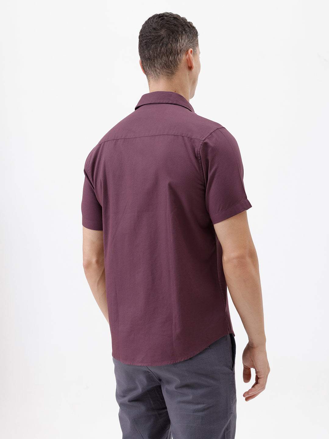 A man modeling a short-sleeve button-up shirt in a deep plum purple color, paired with gray trousers, against a plain white studio background.