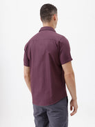 A man modeling a short-sleeve button-up shirt in a deep plum purple color, paired with gray trousers, against a plain white studio background.