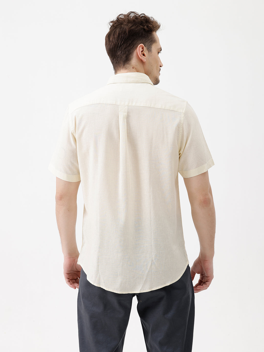 A man wearing a light cream-colored short-sleeve linen button-up shirt with a chest pocket, paired with dark trousers, against a plain white studio background.