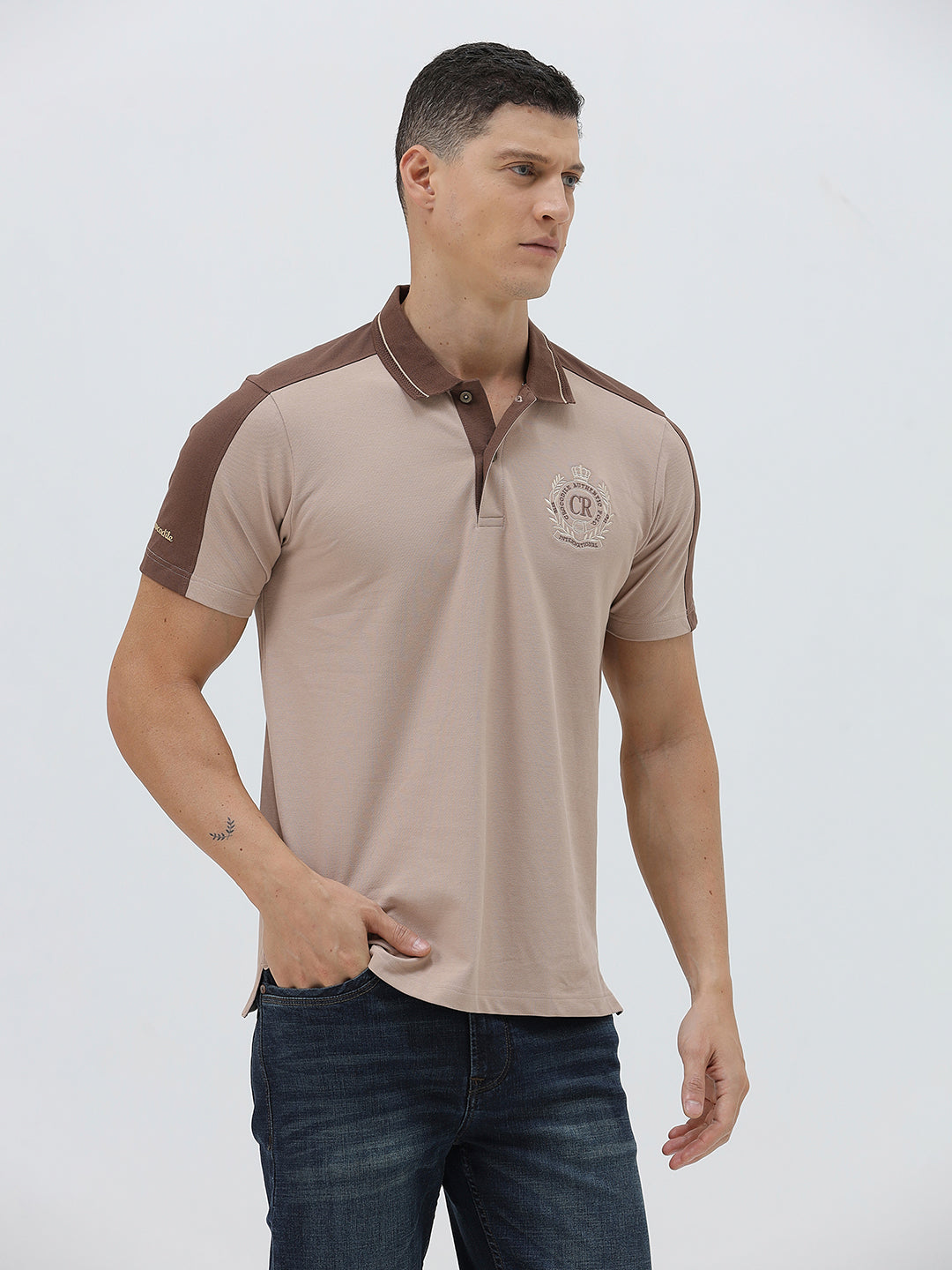 A man modeling a short-sleeve polo shirt in beige with brown shoulder panels and collar trim, paired with dark blue jeans, against a light gray studio background.