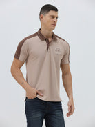 A man modeling a short-sleeve polo shirt in beige with brown shoulder panels and collar trim, paired with dark blue jeans, against a light gray studio background.