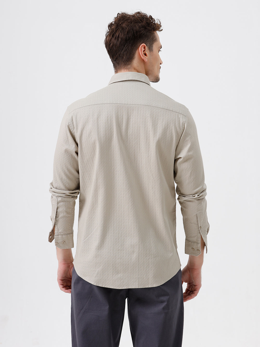 A man wearing a light beige long-sleeve button-down shirt with a chest pocket, paired with dark gray trousers, standing against a plain white studio background.