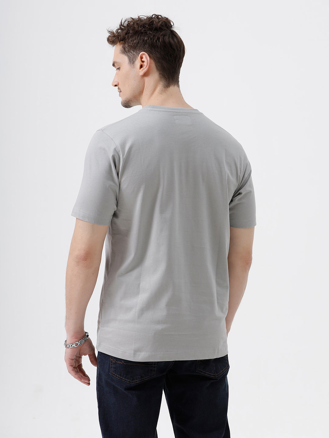 A man wearing a light gray short-sleeve crewneck t-shirt with a subtle tonal chest logo, paired with dark jeans, standing against a light gray studio background.