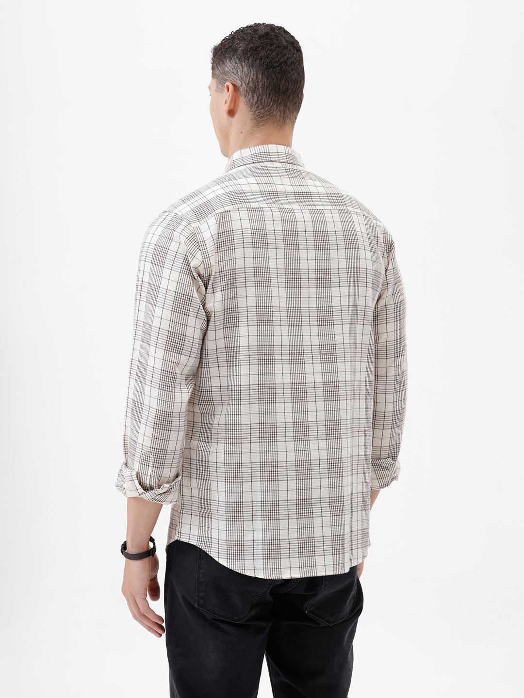 A man wearing a long-sleeve button-up plaid shirt in beige, white, and gray, paired with dark pants, standing against a plain white studio background.