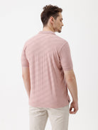A man wearing a dusty pink short-sleeve polo shirt with a chest pocket, paired with light beige trousers, standing against a plain white studio background.