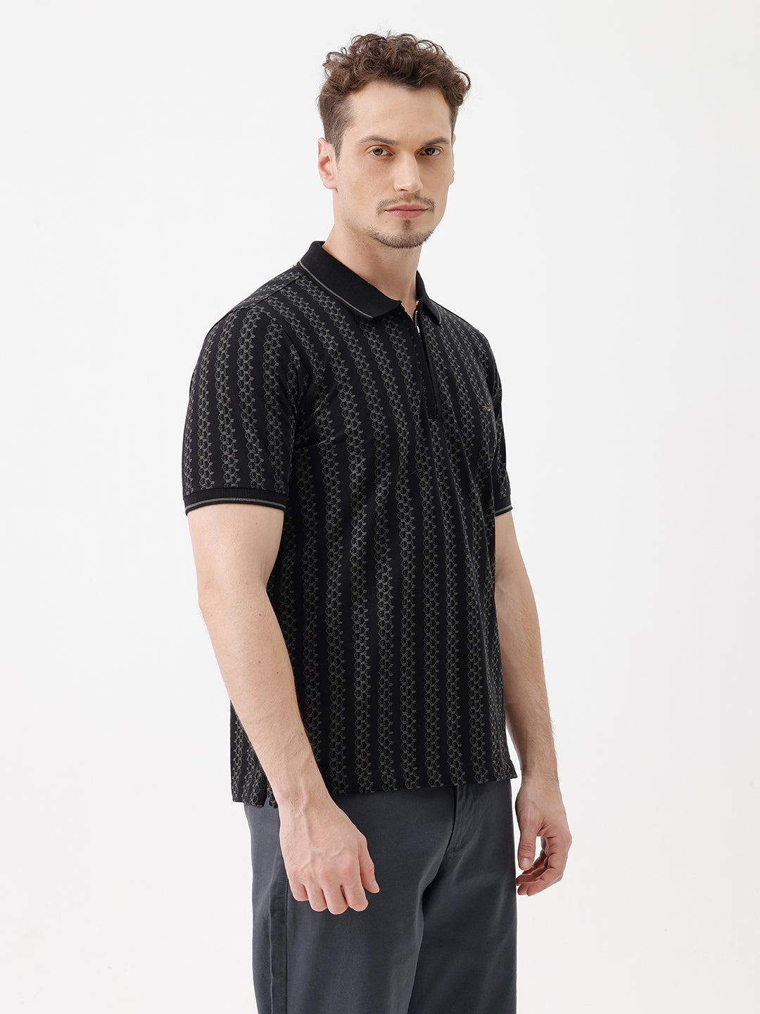 A man wearing a black short-sleeve polo shirt with a subtle geometric pattern, paired with dark gray trousers, standing against a plain white studio background.