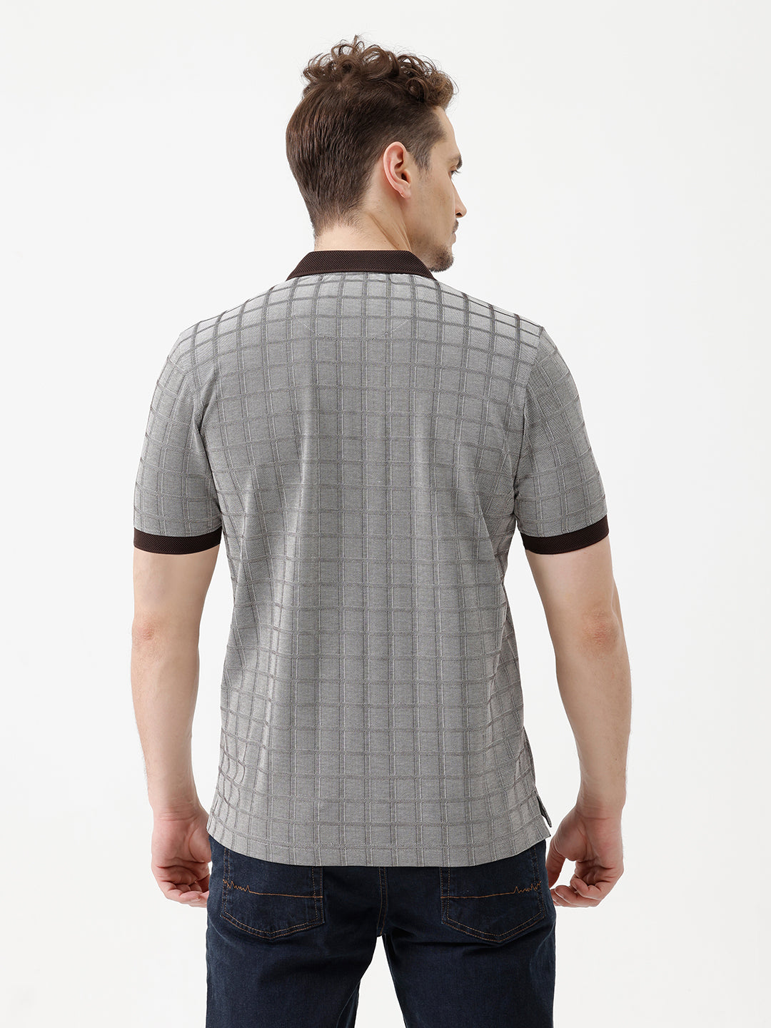 A man wearing a gray short-sleeve polo shirt with a subtle grid pattern and dark brown collar and sleeve trim, paired with dark blue jeans, against a plain white studio background.