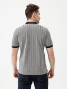 A man wearing a gray short-sleeve polo shirt with a subtle grid pattern and dark brown collar and sleeve trim, paired with dark blue jeans, against a plain white studio background.