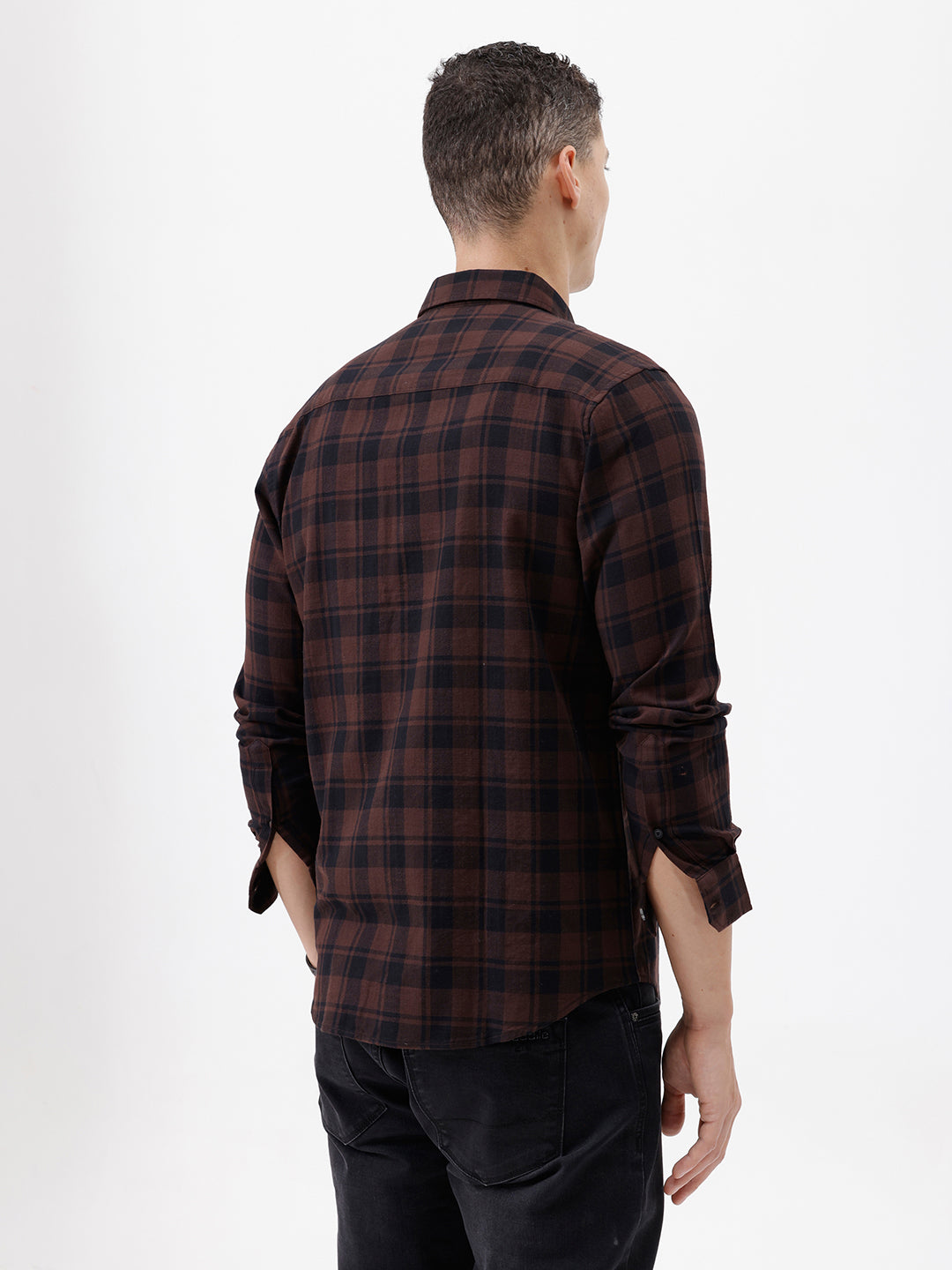 A man wearing a dark brown and black plaid long-sleeve button-up shirt, paired with dark pants, standing against a plain light gray background.