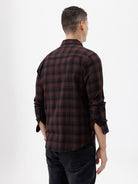 A man wearing a dark brown and black plaid long-sleeve button-up shirt, paired with dark pants, standing against a plain light gray background.