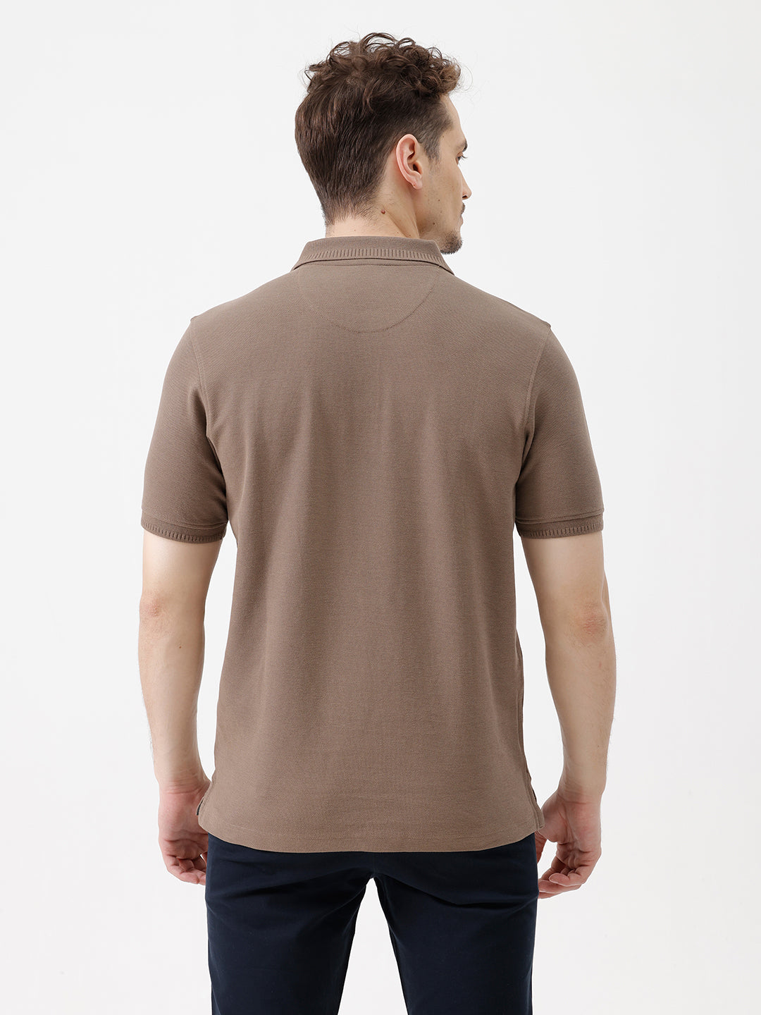 A man wearing a brown short-sleeve polo shirt with dark navy pants, standing against a plain light gray studio background.