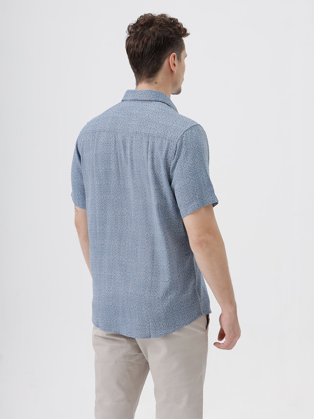 A man wearing a short-sleeve button-up shirt with a fine blue and white geometric pattern, paired with light beige trousers, standing against a plain light gray background.