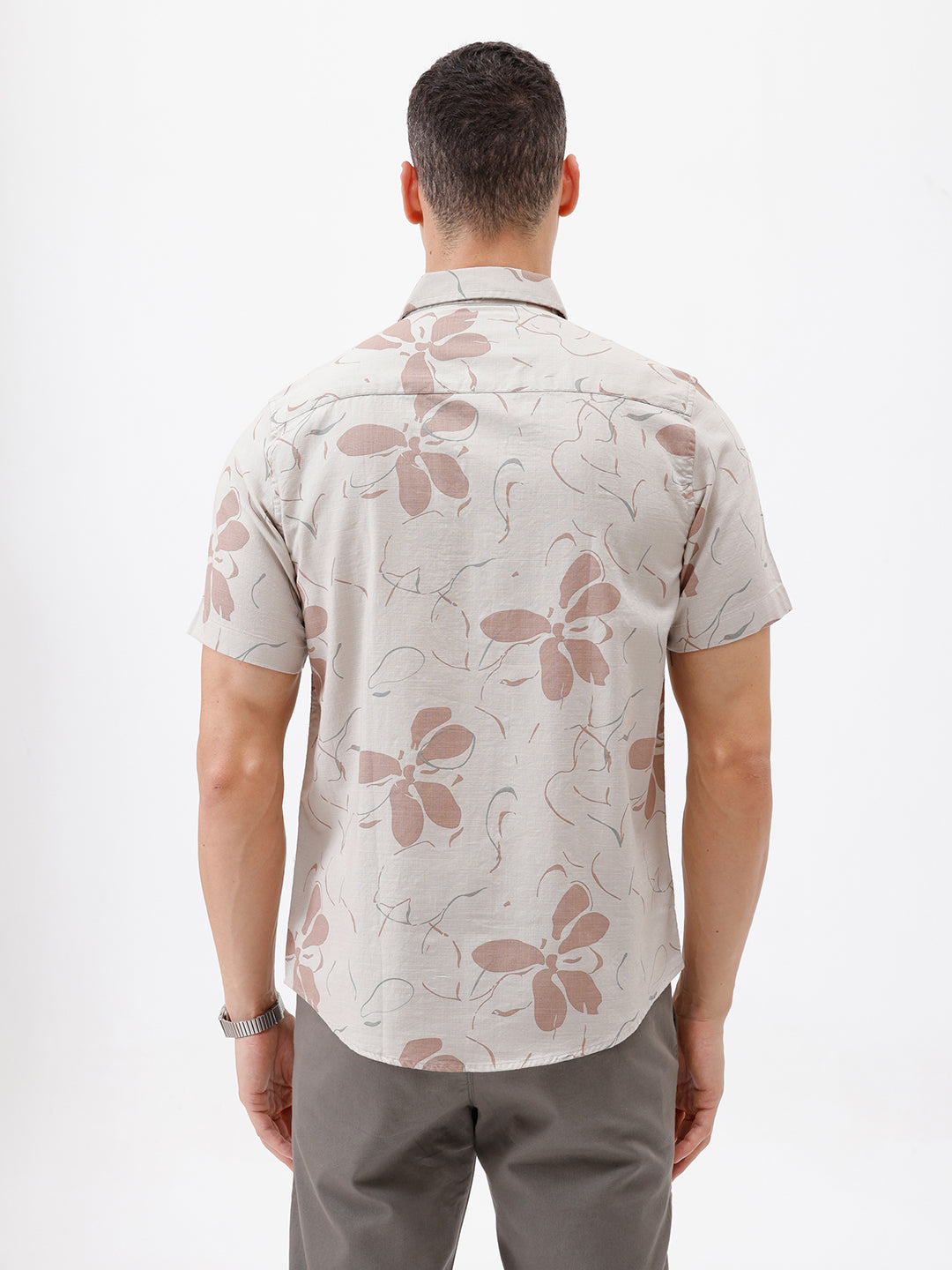 A man wearing a short-sleeve button-up shirt with a beige and brown floral pattern, paired with gray trousers, standing against a plain white background.
